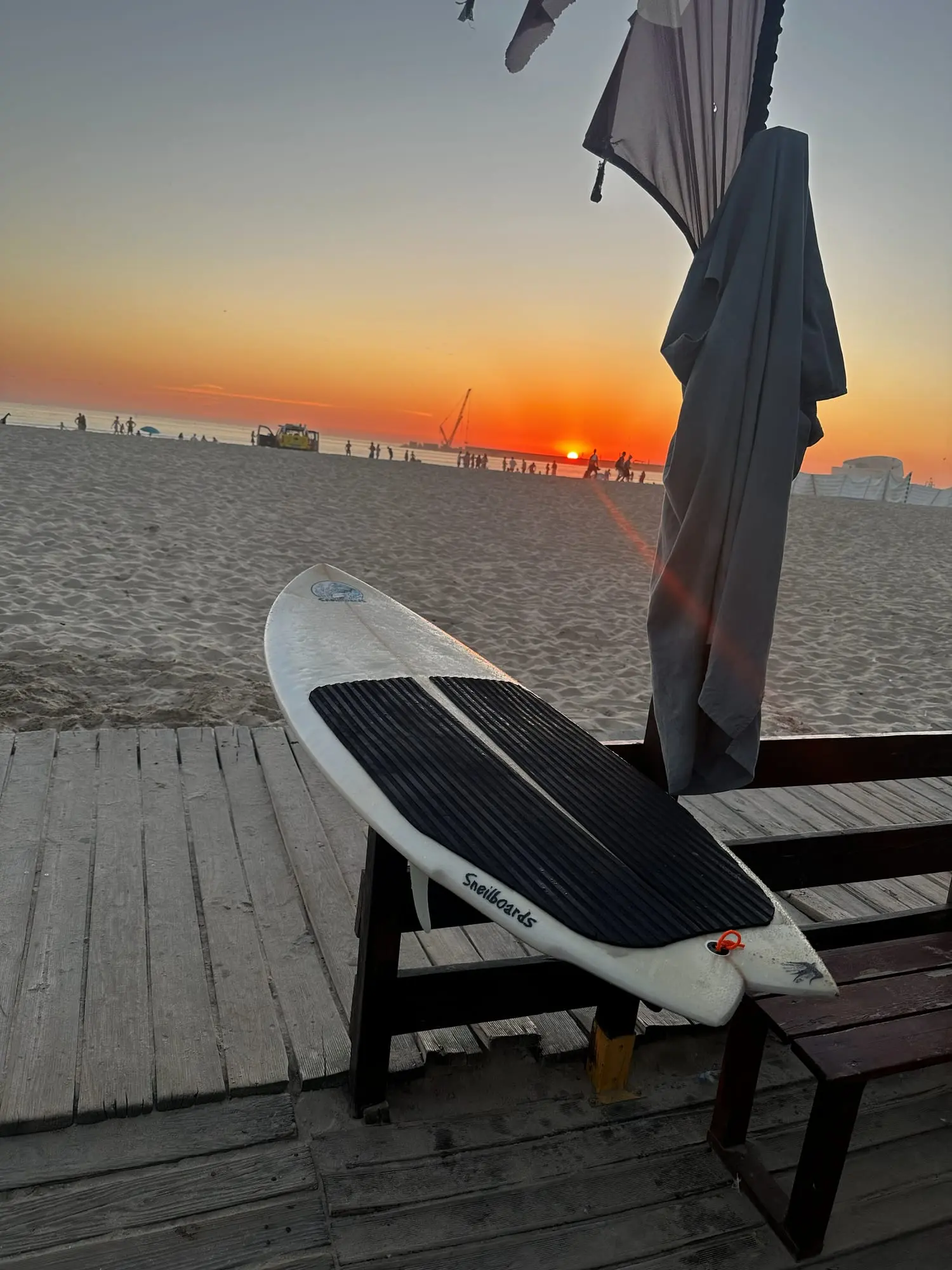 A surfboard leaning on a wooden bench on a beach boardwalk at sunset, with a wetsuit hanging beside it