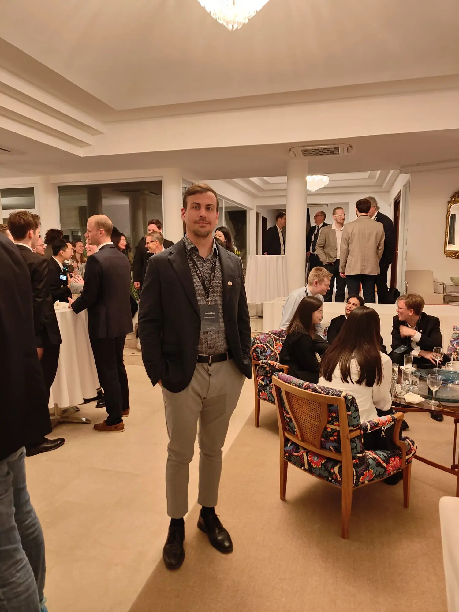 Dominik standing in a blazer and lanyard while guests talk at tables behind him