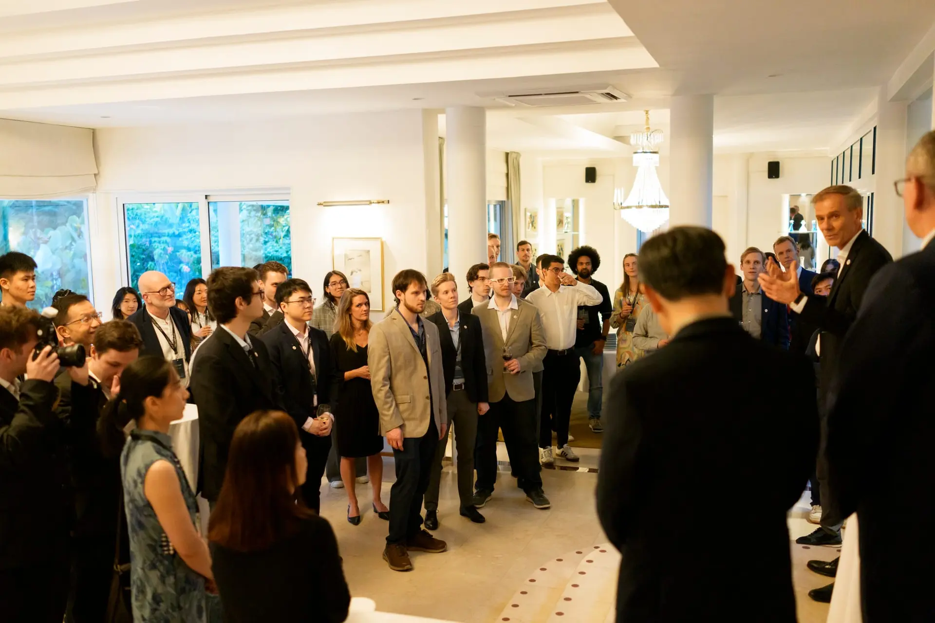 WASP students and guests standing in a white reception room listening to the Ambassador