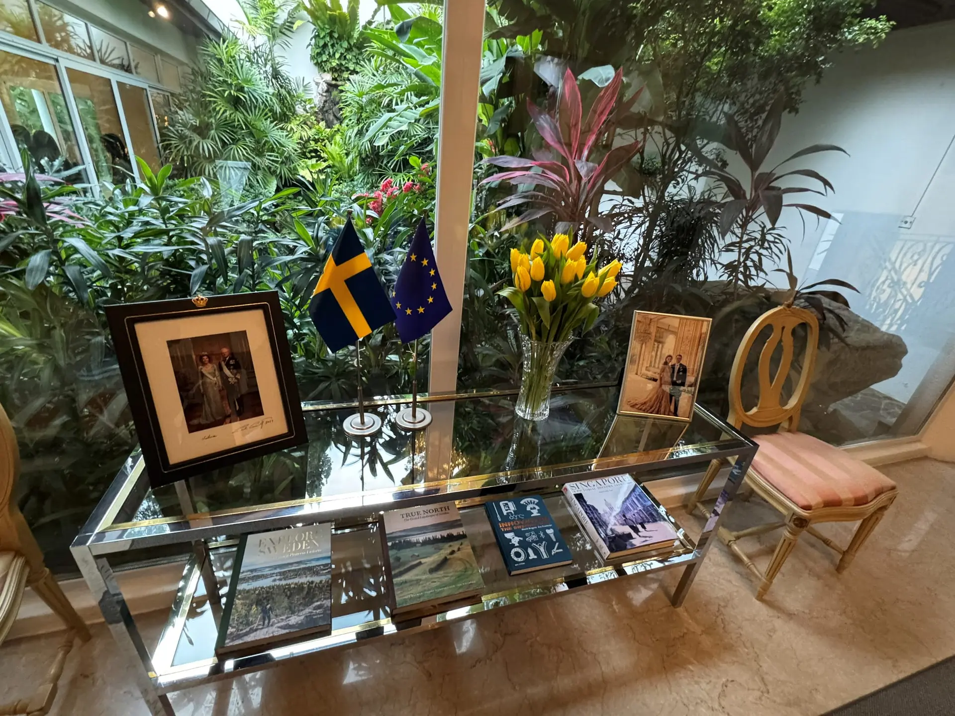 A glass table with Swedish and EU flags, yellow tulips, framed portraits and books about Sweden and Singapore, in front of a tropical garden