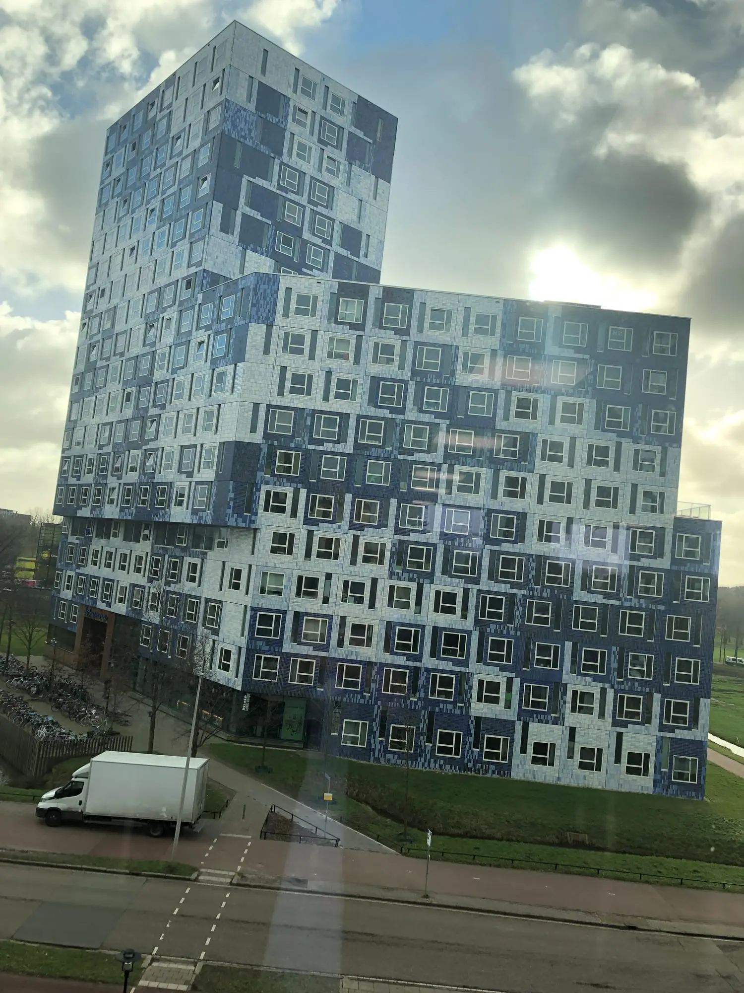 A large residential block tiled in blue and white squares, seen through a window above a road and a lawn