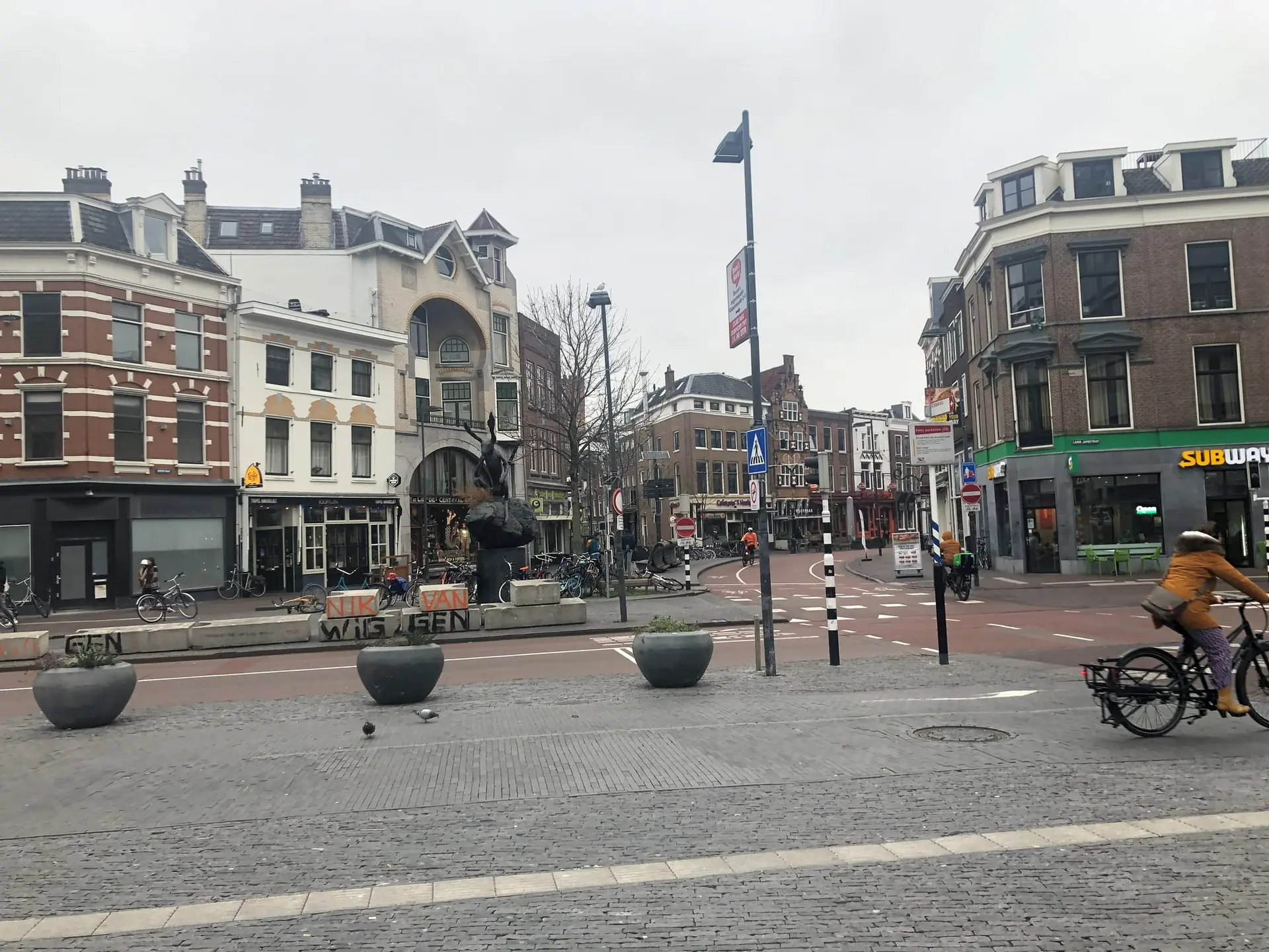 A square in Utrecht's city centre on a grey day, with brick town houses, a dark statue, large plant pots and a cyclist on a red bike lane