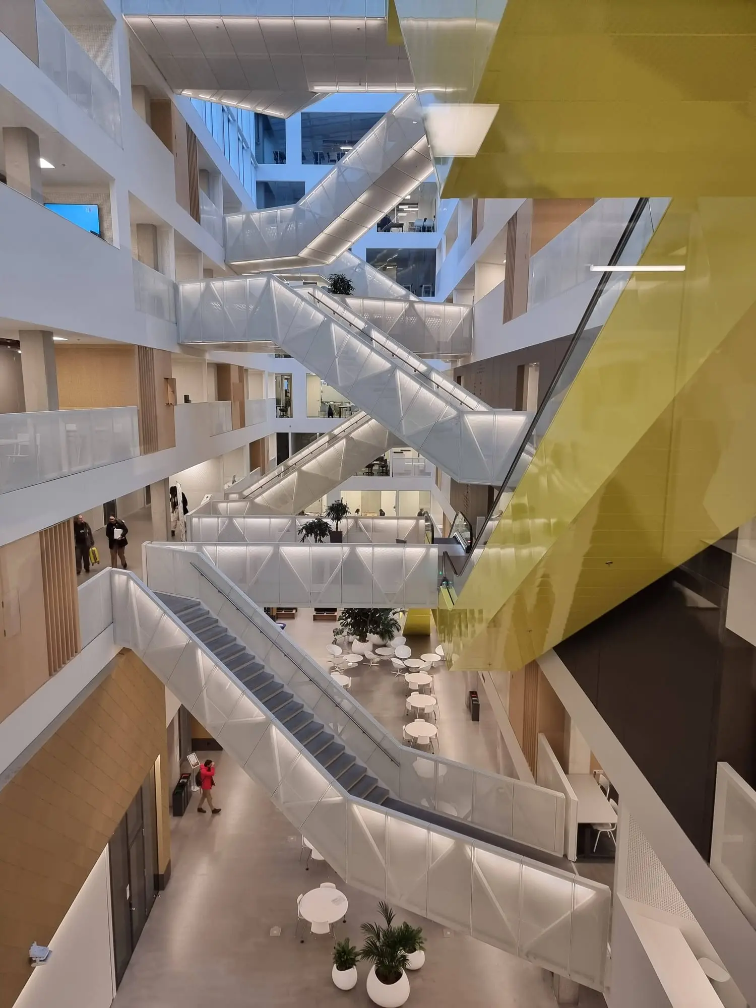 A tall atrium with white staircases crossing between floors, a yellow escalator on the right and white tables and chairs on the ground floor