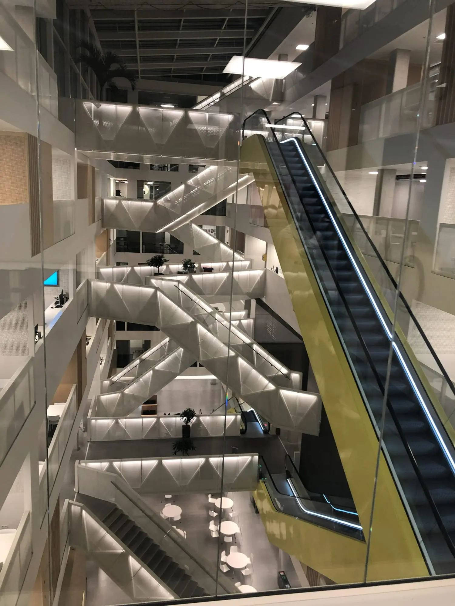 A long escalator in a yellow casing rising through an atrium of white staircases, seen through a glass wall