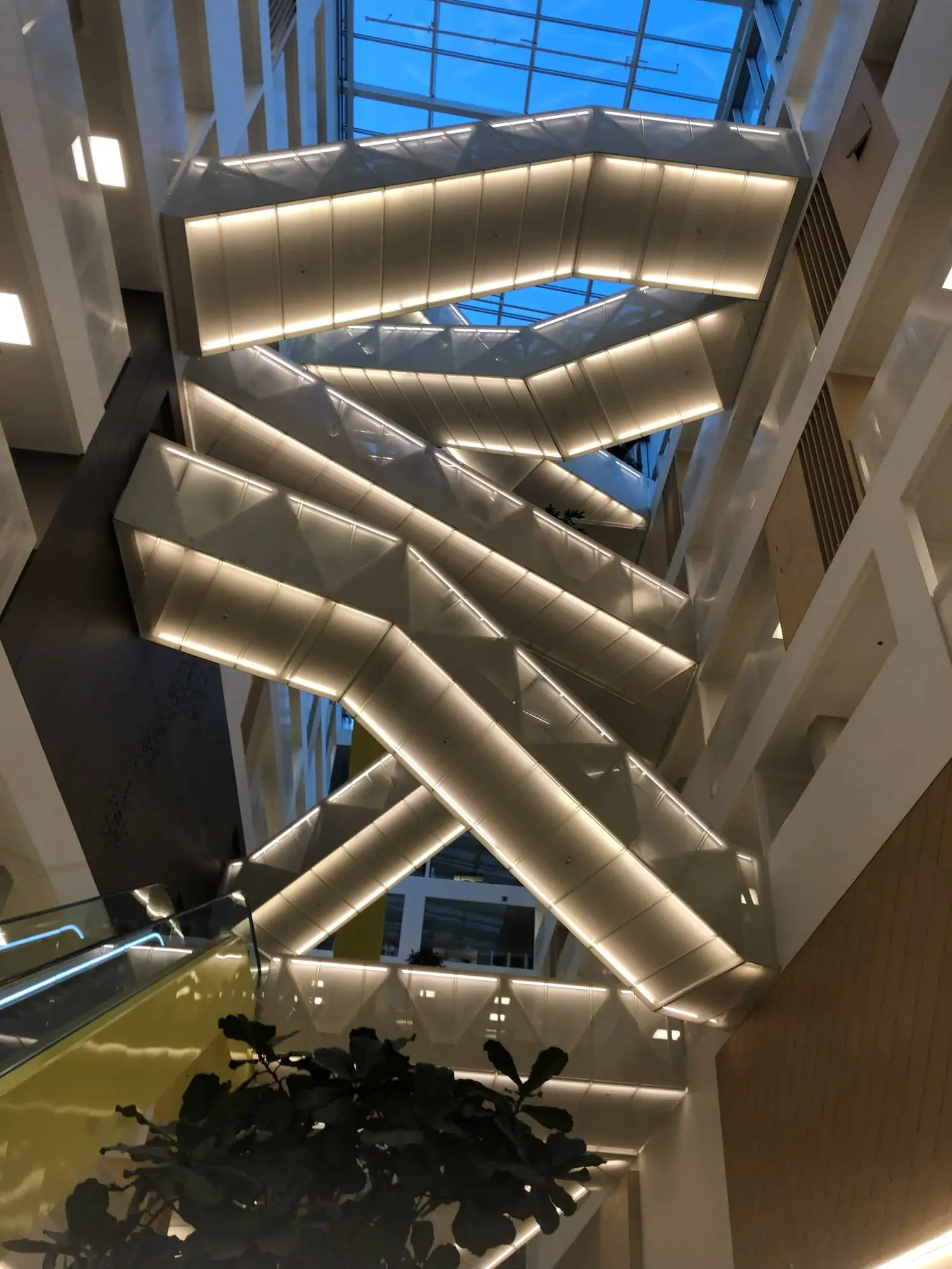 Looking up through a tall atrium at white zigzag staircases lit from below, under a glass roof