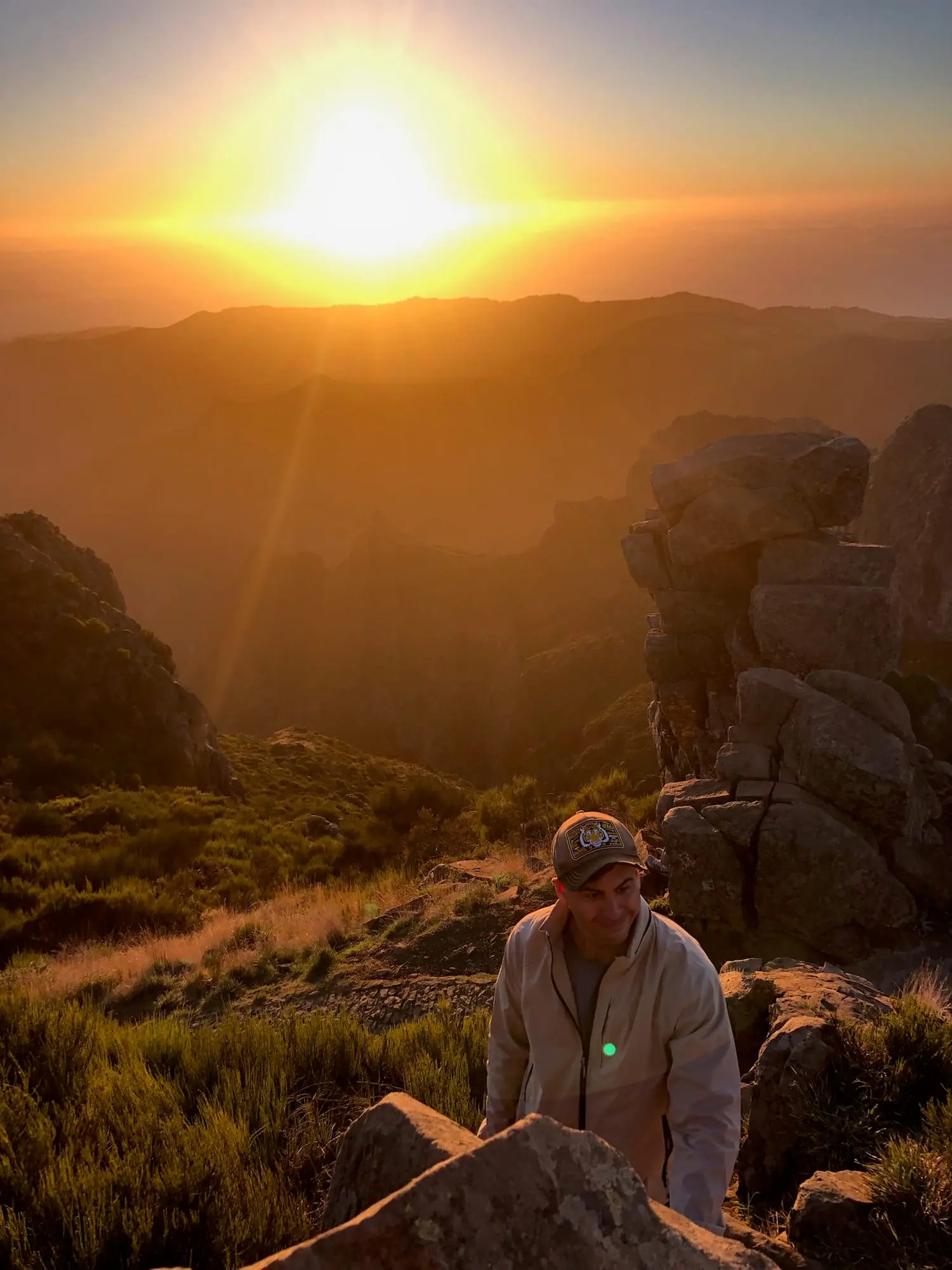 A hiker in a cap climbing between rocks as the sun rises over the mountain ridges of Madeira