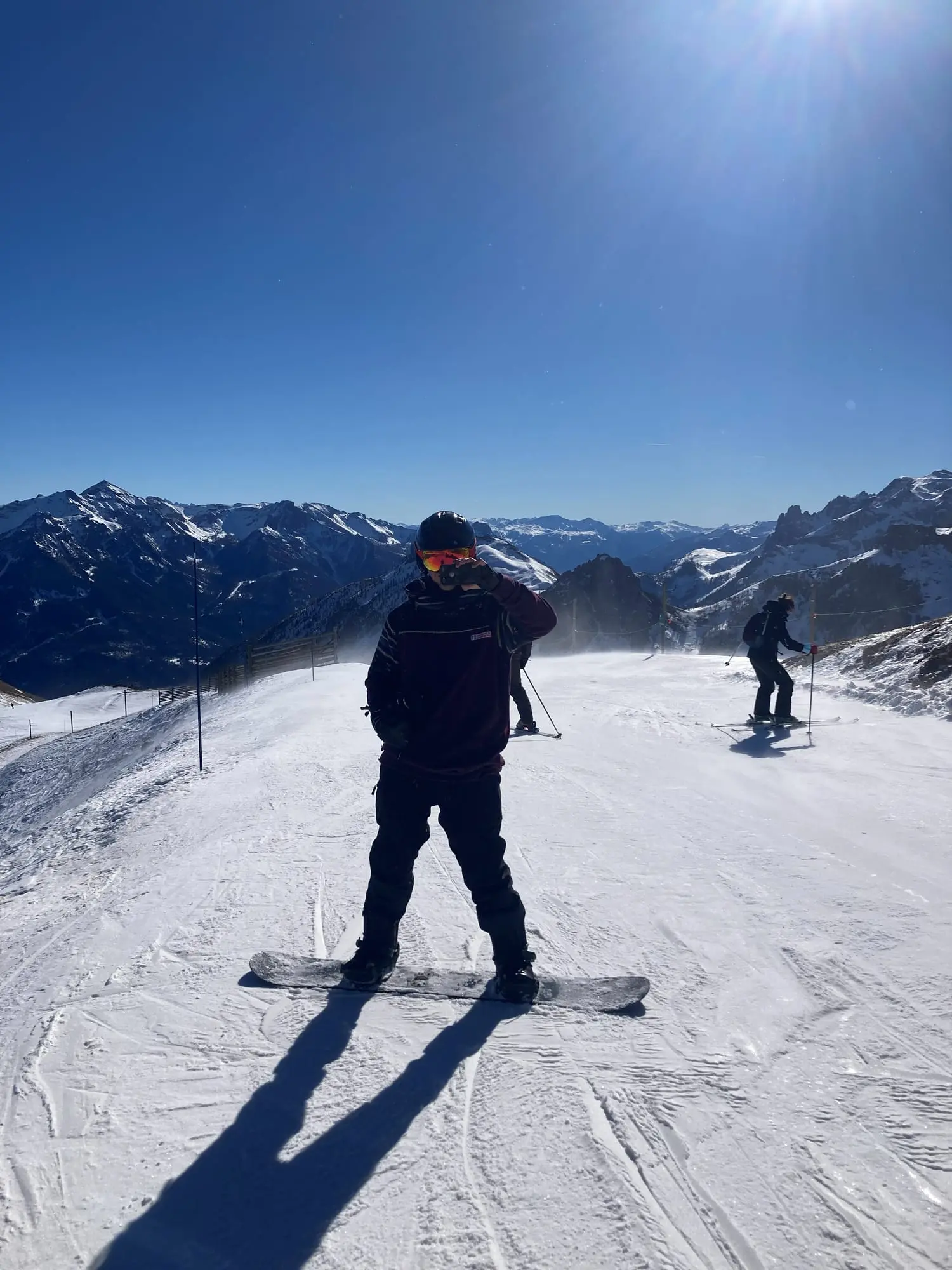 A snowboarder standing on a groomed piste under a clear blue sky, with snowy mountain ridges behind