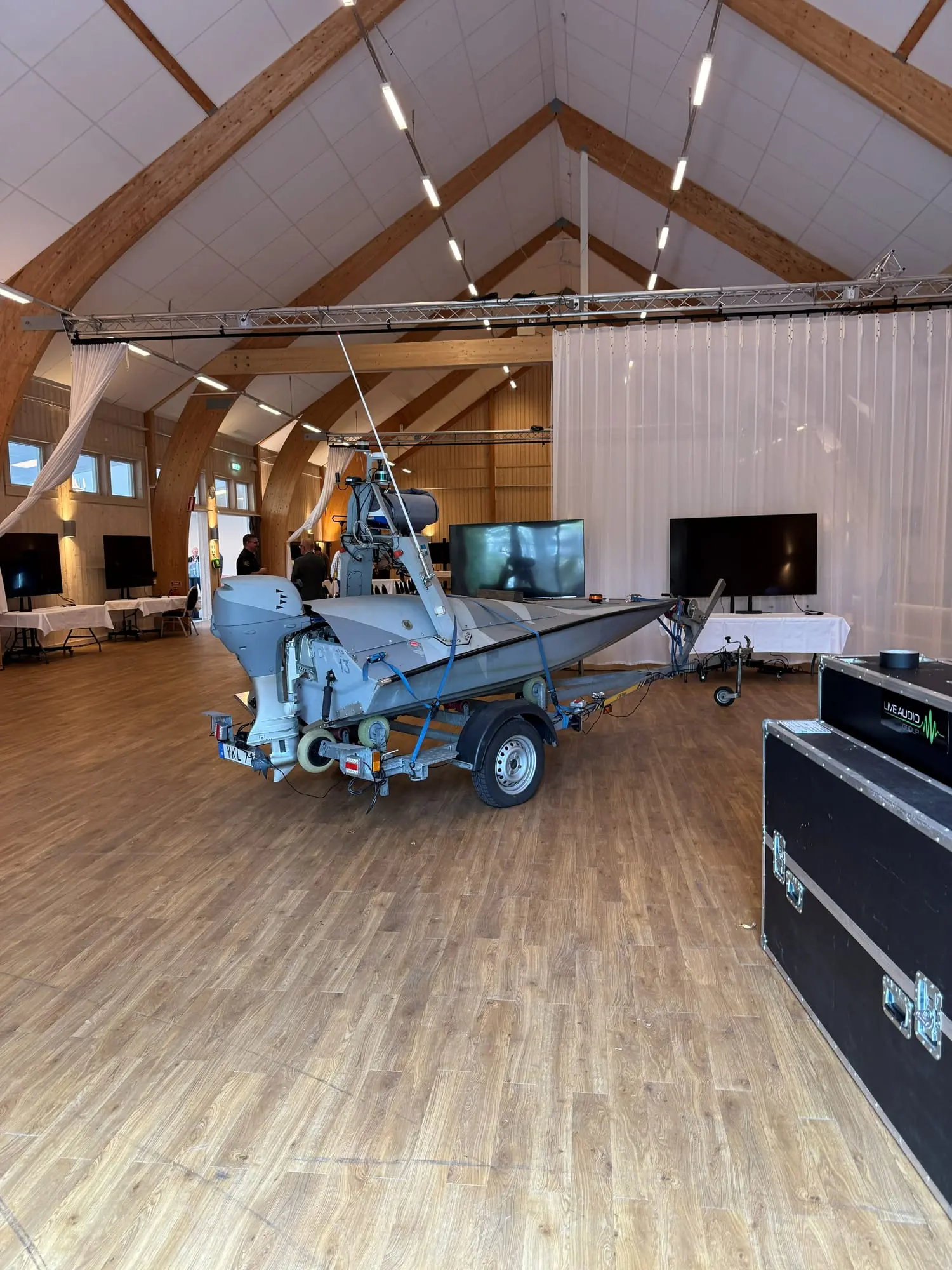 A small research boat with a sensor mast on a trailer in a large exhibition hall with display screens