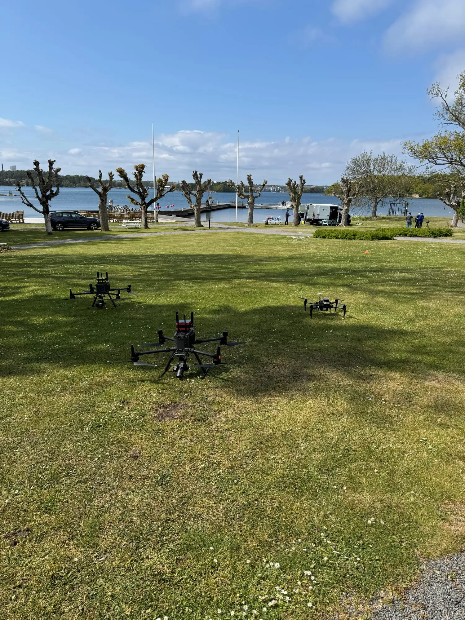Three quadcopter drones on a lawn by the water, with a jetty and a van in the background