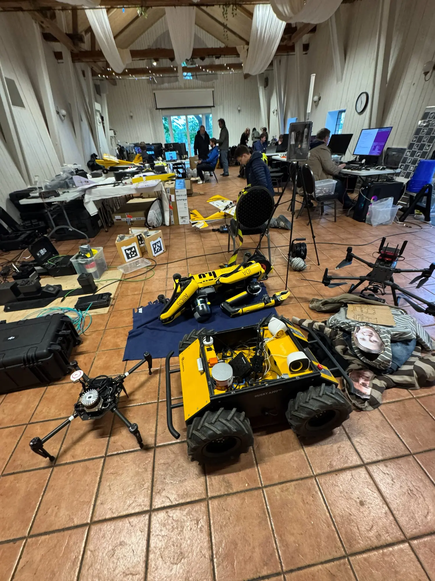 A hall full of robots and equipment: a legged robot, a wheeled ground robot, drones and fixed-wing aircraft, with researchers working at laptops