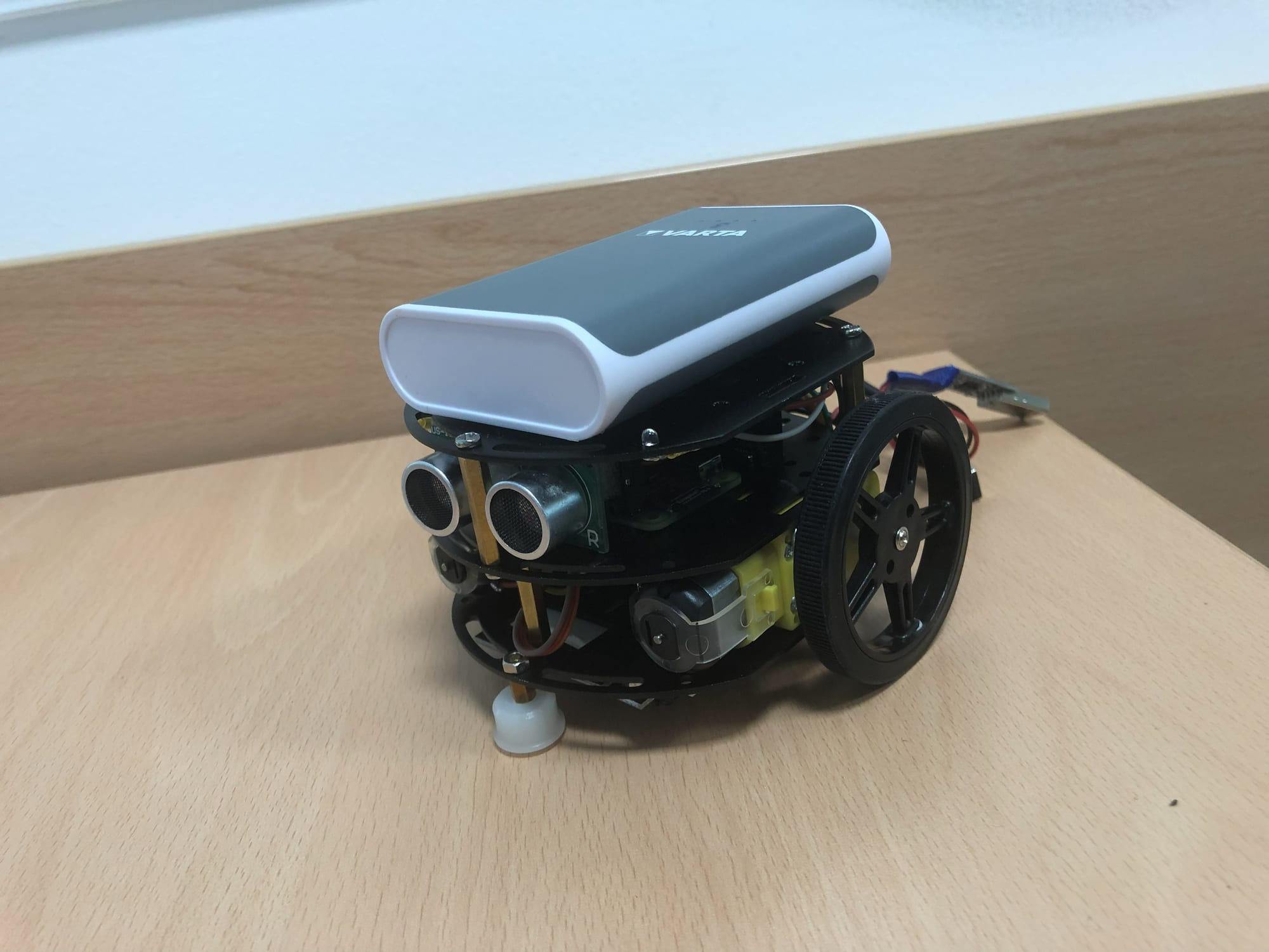 Close-up of one robot car: stacked chassis plates, an ultrasonic rangefinder at the front, two wheels and a power bank on top