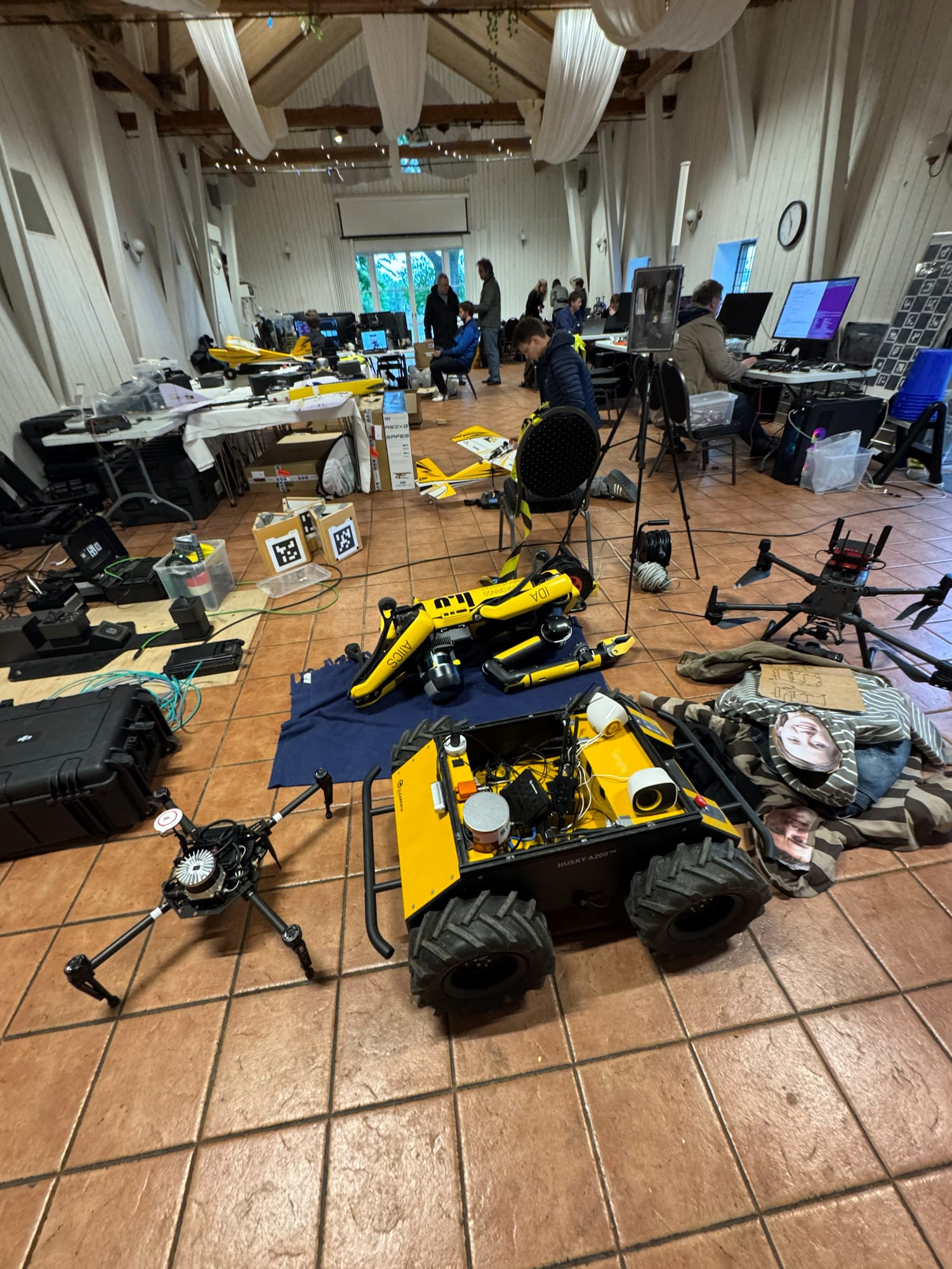 A hall full of robots and equipment: a legged robot, a wheeled ground robot, drones and fixed-wing aircraft, with researchers working at laptops